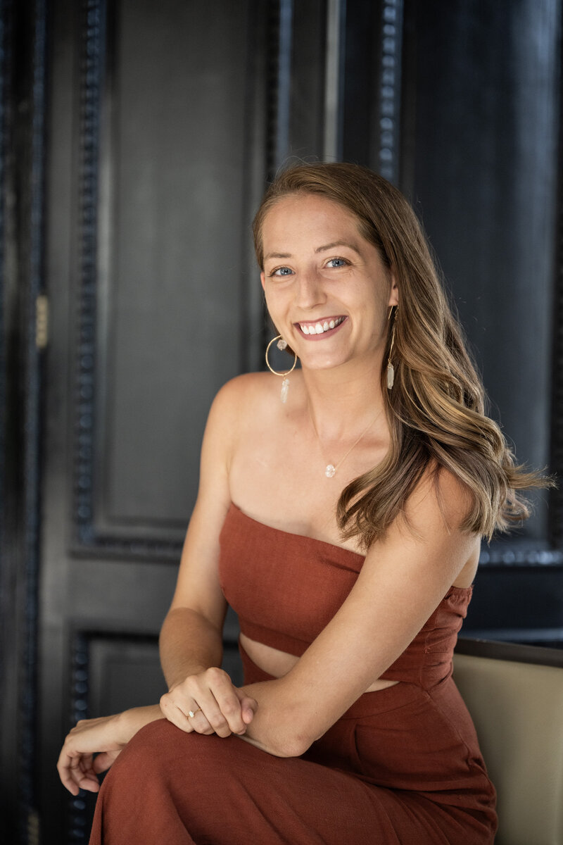 Katie sits on a chair in a jumpsuit smiling at the camera.