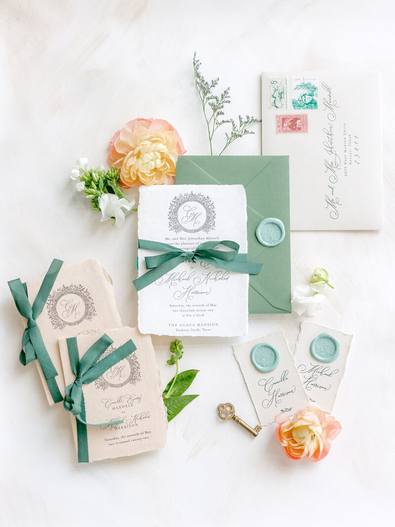Flat lay of white and green wedding invitation suite with green ribbons, wax seals, and orange flowers on a white background