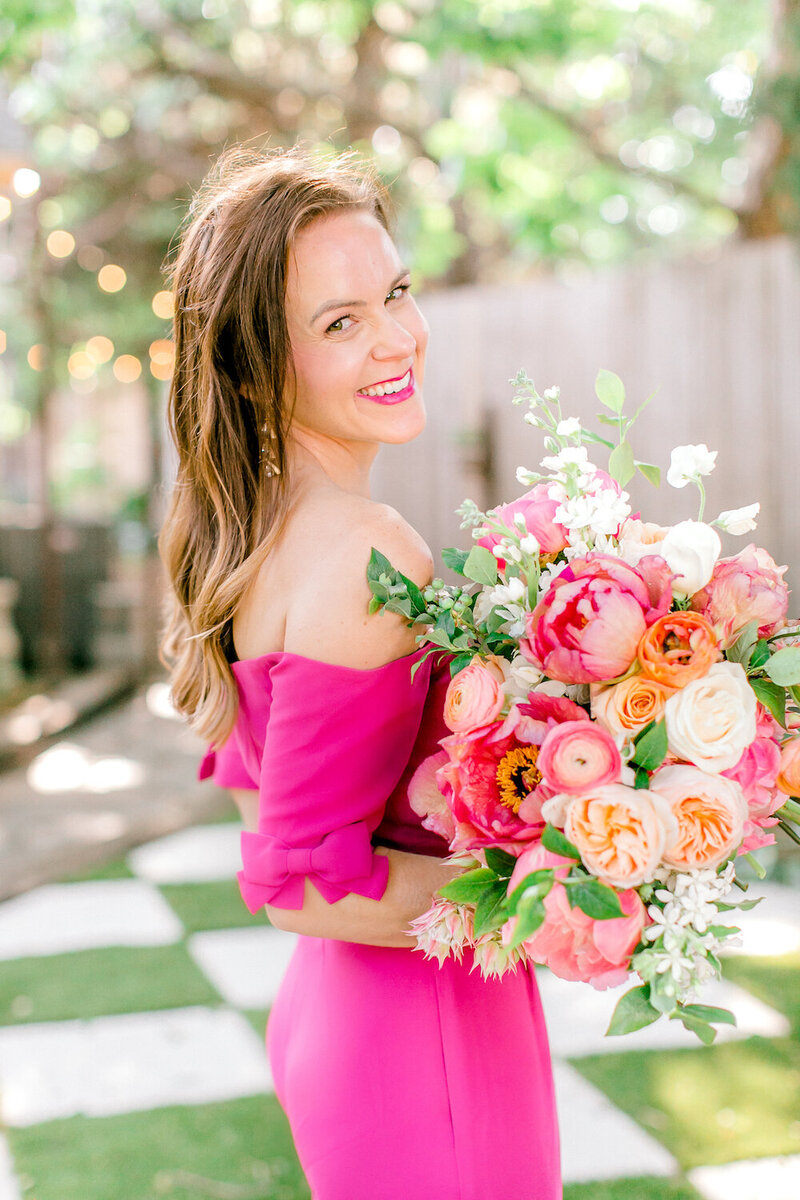 Meg in a hot pink dress, holding a colorful bouquet and smiling at the camera.