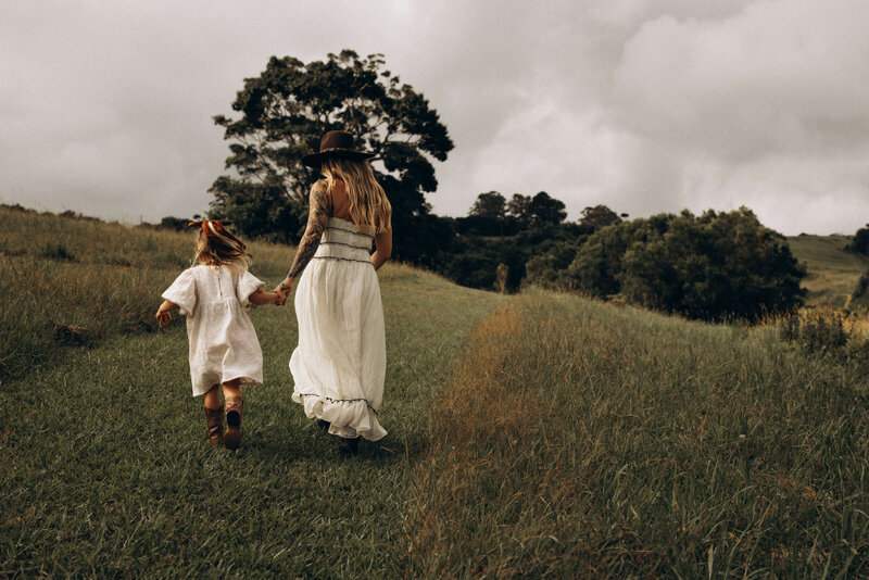 A mother and daughter walk hand-in-hand through an open grassy field under a cloudy sky, their dresses flowing as they move forward together.