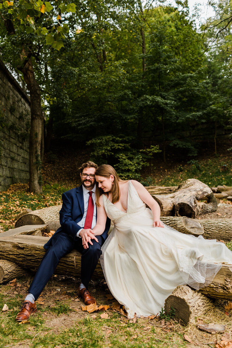 A bride and groom sitting on logs in an autumn park holding hands