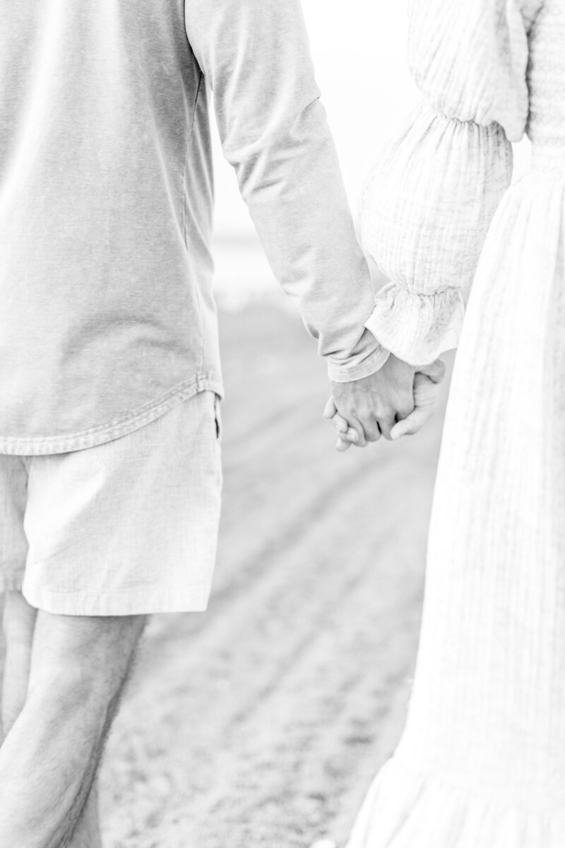 Black and white image of a couple holding hands while walking barefoot along the shoreline — intimate Kansas City maternity portrait focused on connection.