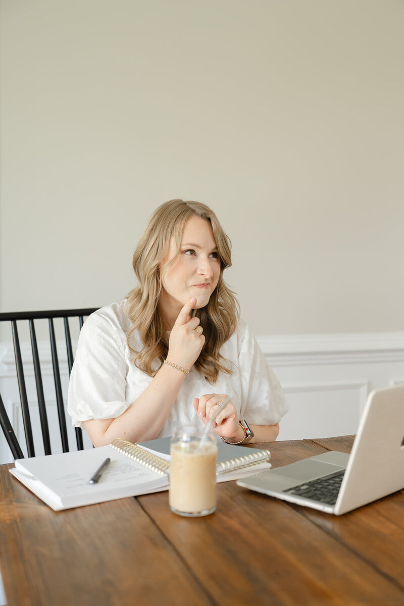 Woman in a white blouse touching her pointer finger to her chin and thinking while seated at a wooden farm table with a Macbook and iced coffee in front of her