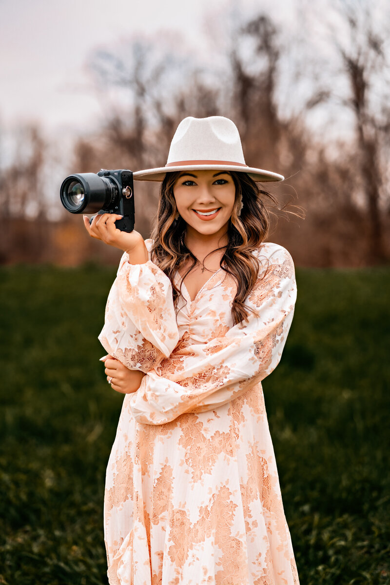 Emily Belanger: Elopement Photographer holding a camera and smiling while standing in a field with the Adirondack mountain range in the background