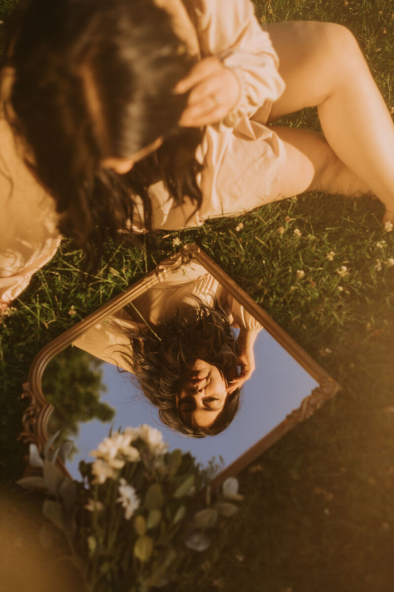 Woman with long brown hair in a tan dress sits outdoors in grass and stares into mirror admiring her reflection
