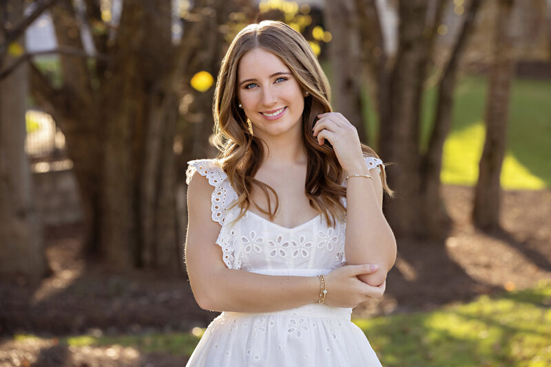 Beautiful high school senior in a white dress standing in near backlit trees and flowers at Anne Springs Close Greenway, surrounded by soft autumn colors