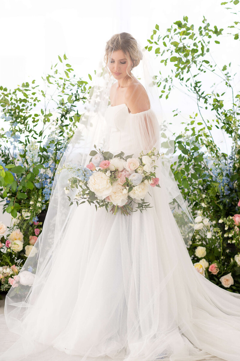 a bride holding a white, pink and blue bouquet, standing under her cathedral length veil in her floral ceremony site.  Captured indoors by Ottawa wedding photographer JEMMAN Photography