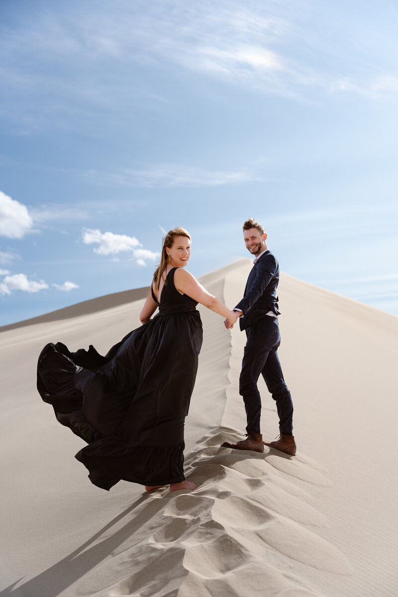 A couple holds hands on a sand dune and looks back at the camera smiling