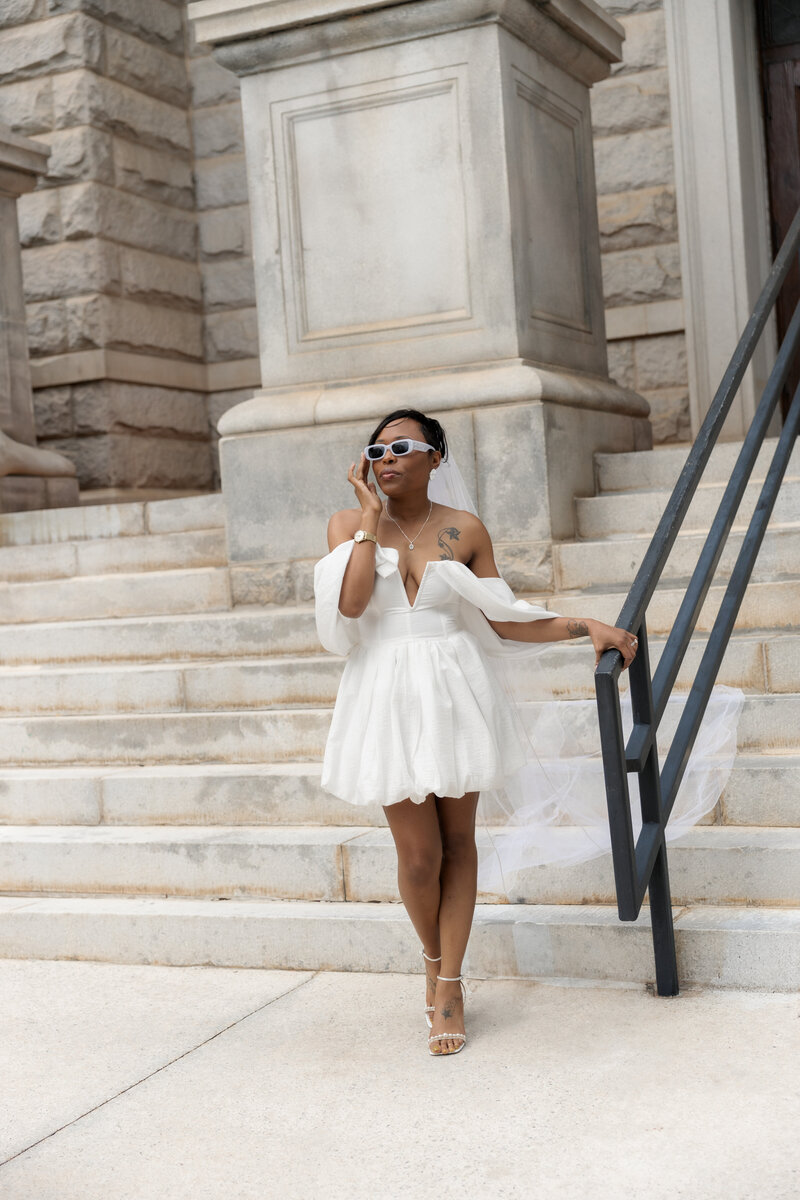 A chic bride in a short white dress and veil, wearing sunglasses and standing confidently on the steps of an Atlanta courthouse.