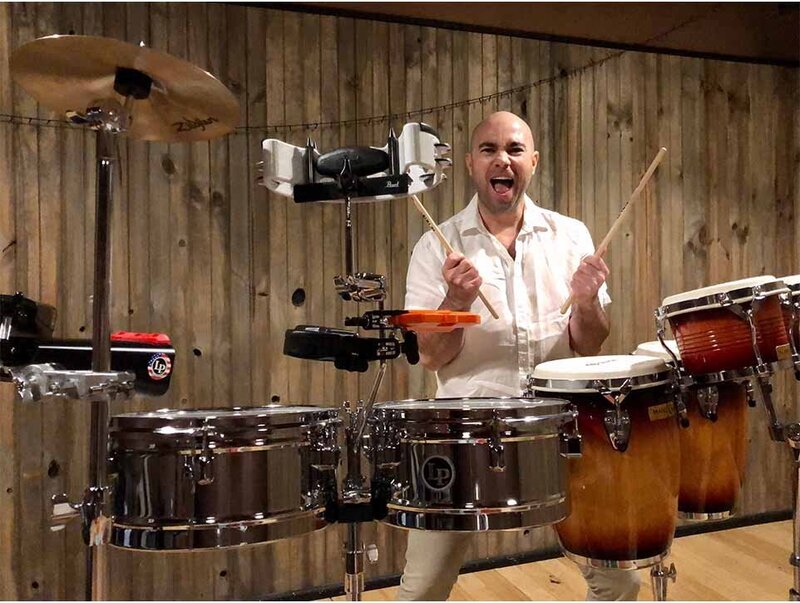 Energetic percussionist holding drumsticks mid-performance surrounded by timbales, congas, and cymbals against wooden paneling.
