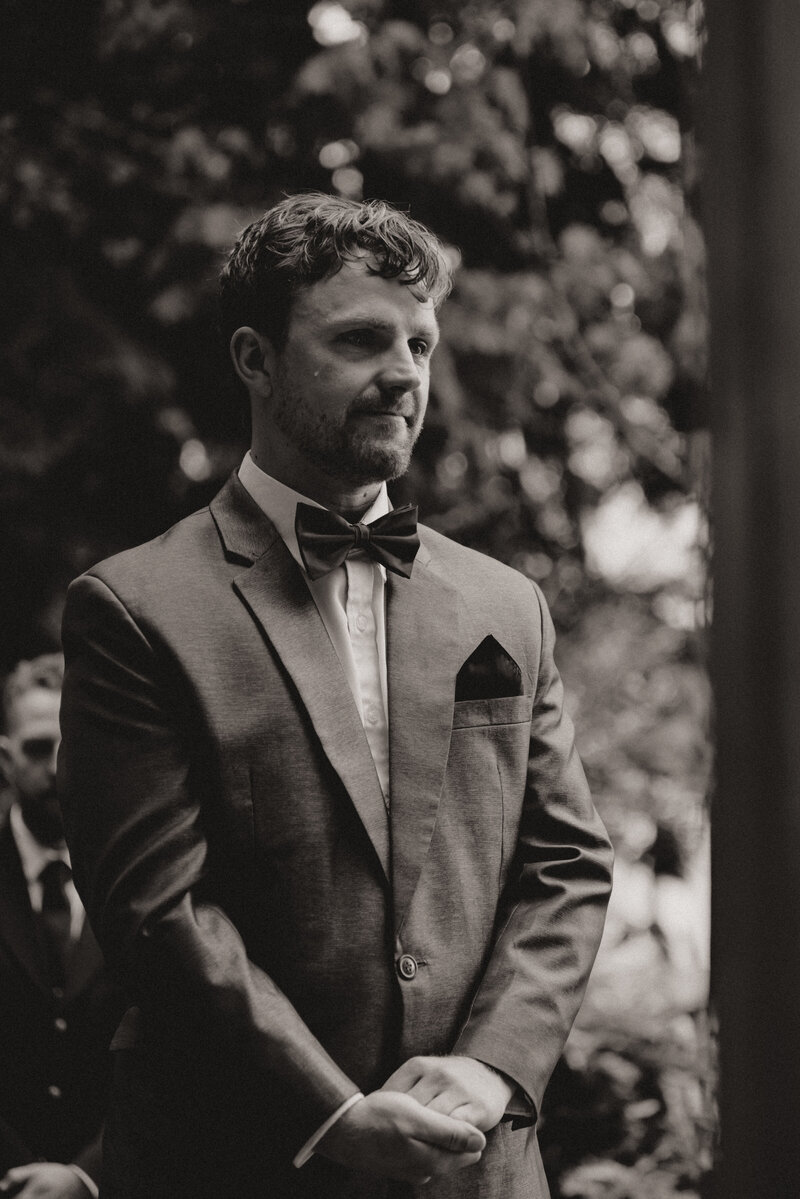 A black ablack and white photo of a groom crying a tear while his bride walks down the aislend white photo of a bride placing a wedding band on the hand of her groom. The close-up photo shows off their wedding rings, the sleeves of the groom's suit, manicured nails of the bride, and her black cross tattoo on her inner wrist.