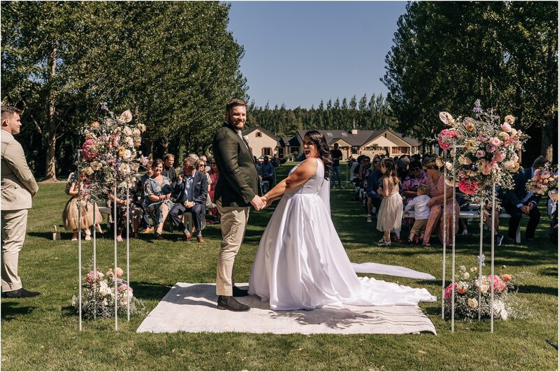 bride and groom at ceremony on lawn with guests behind floral plinths pink orange ellies belly