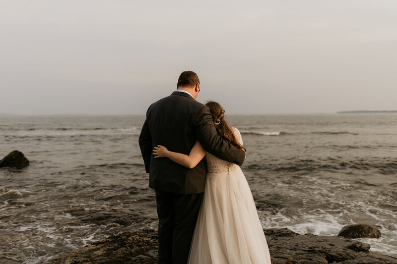 A couple embraces in a hug overlooking the coast of Acadia National Park