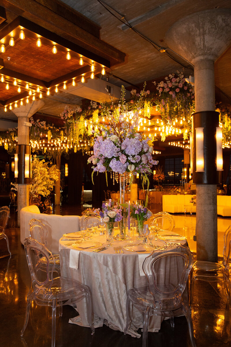 Round table draped in champagne linen with tall lavender florals, acrylic ghost chairs, and warm hanging lights at an indoor reception.