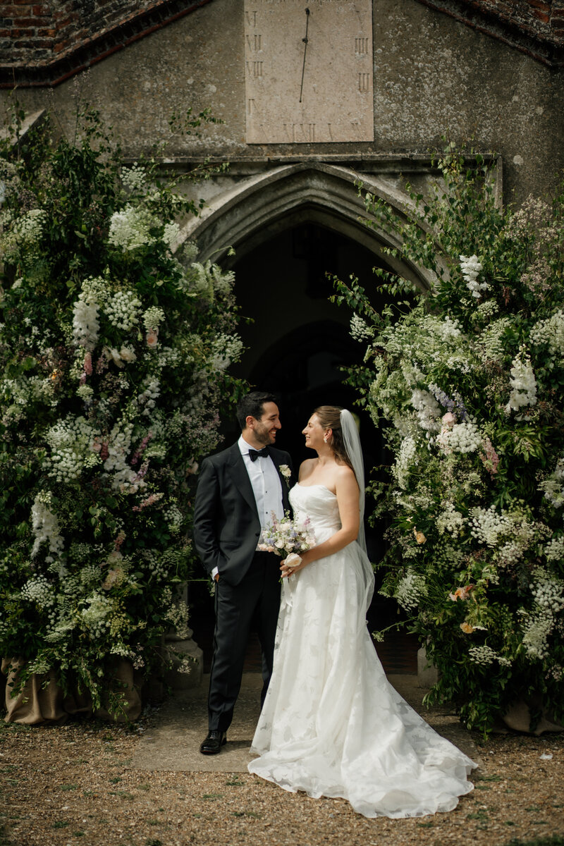 Bride and groom posing in front of flower arch after their ceremony captured by Suffolk Wedding Photographer Lucy of The Unscripted