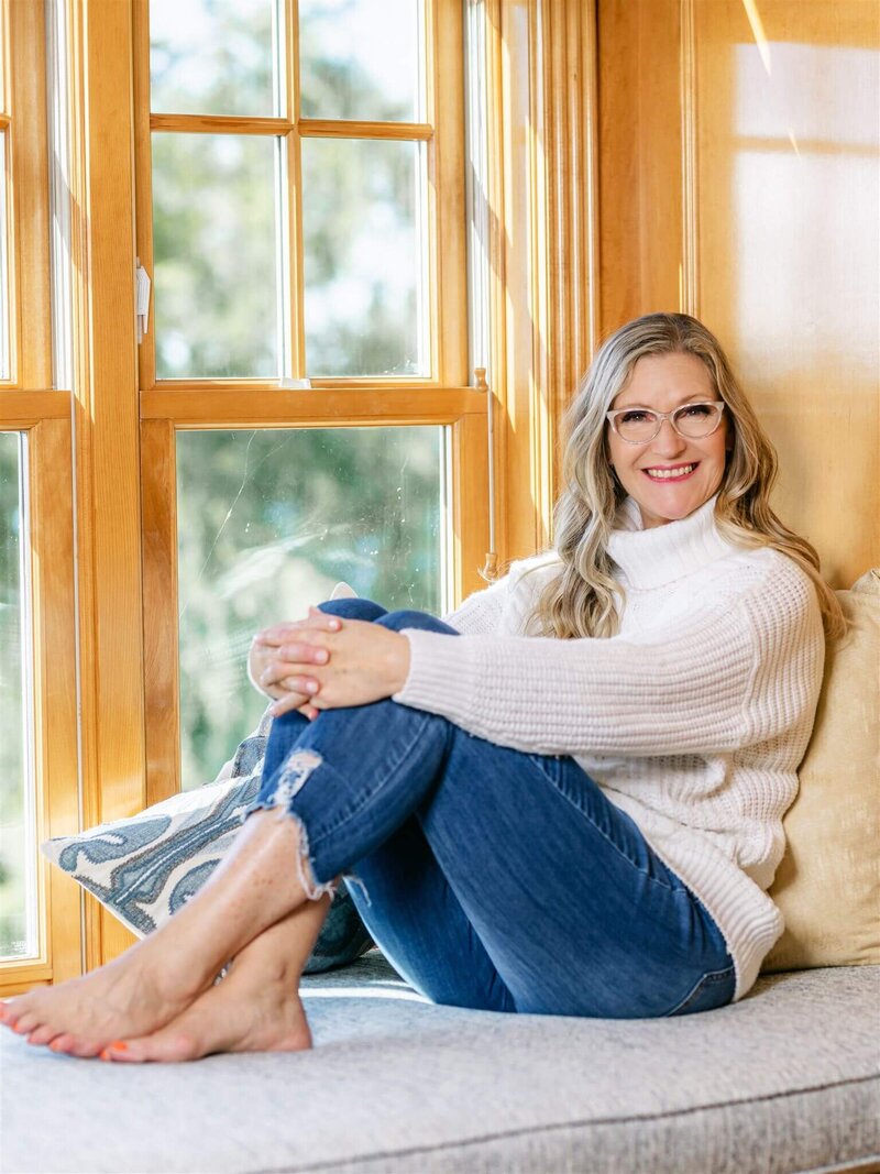 Phillipa Leseberg in a white sweater and jeans sits on a window seat, an open book resting on her lap, smiling with her legs crossed.
