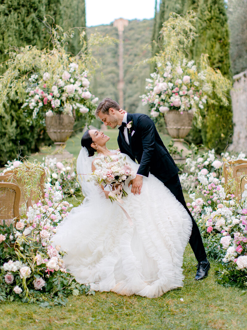 Groom in black tux dips bride in the center of their wedding ceremony aisle surrounded by white and pink roses