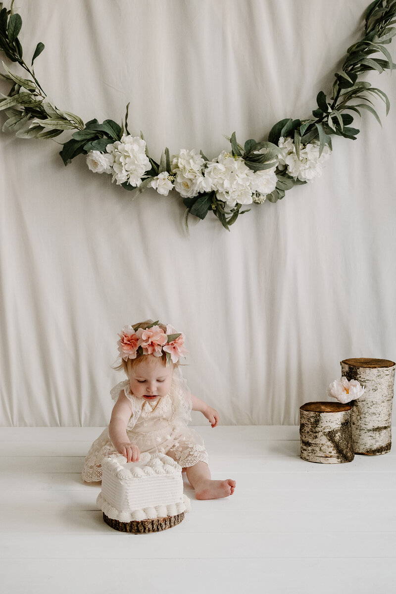 Baby girl in white lace with floral crown sitting behind a white cake in a white studio in Annapolis Maryland photographed by Bethany Simms Photography