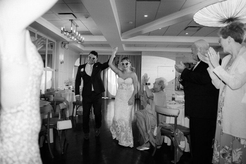 Bride and groom burst into their reception with matching heart shaped glasses on, smiles on their faces and hands raised in the air.
