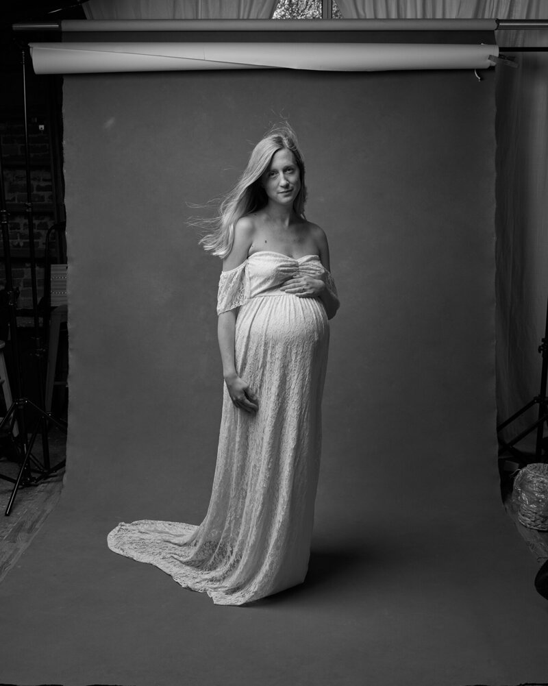 Black and white portrait of a pregnant woman standing gracefully in a flowing, off-the-shoulder lace gown. Her long hair moves gently, and she rests a hand on her belly with a serene, confident expression. The raw studio setting, with an exposed backdrop and natural light, adds to the organic beauty of this unfiltered moment. A powerful, intimate capture from 'THIS IS ME – A Photography Experience by Katie Campbell' in Atlanta, GA, celebrating motherhood and self-expression.