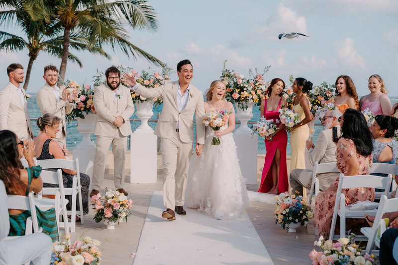 Joyful wedding ceremony exit by the beach, with the bride and groom walking down the aisle hand in hand, smiling and celebrating as guests cheer