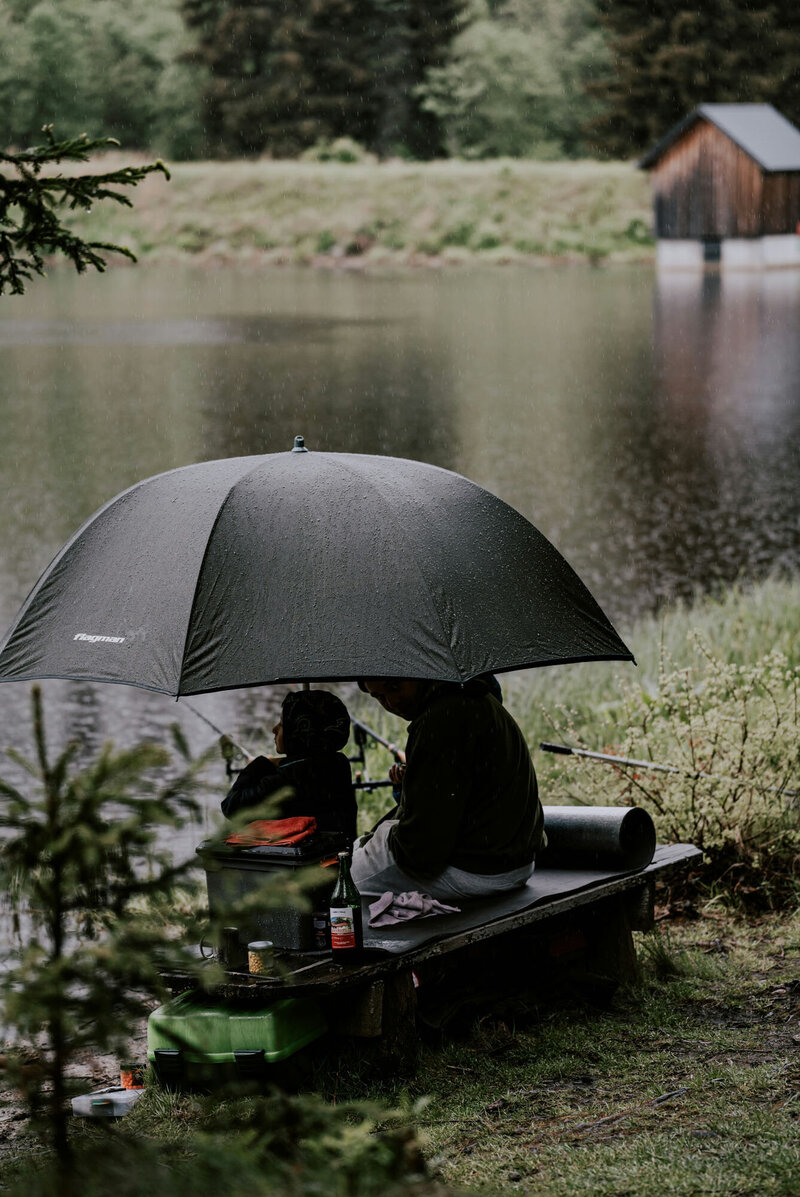 Man is sitting on a bench holding an umbrella over his head while fishing.