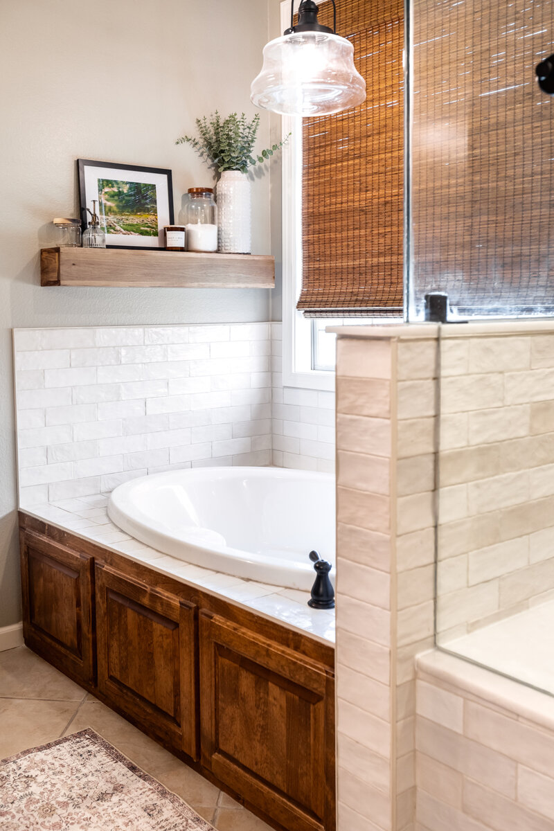 Soaker tub with stained cabinet tub front and tile tub deck and surround with decorative floating shelf with bath decor and large window to let in light.
