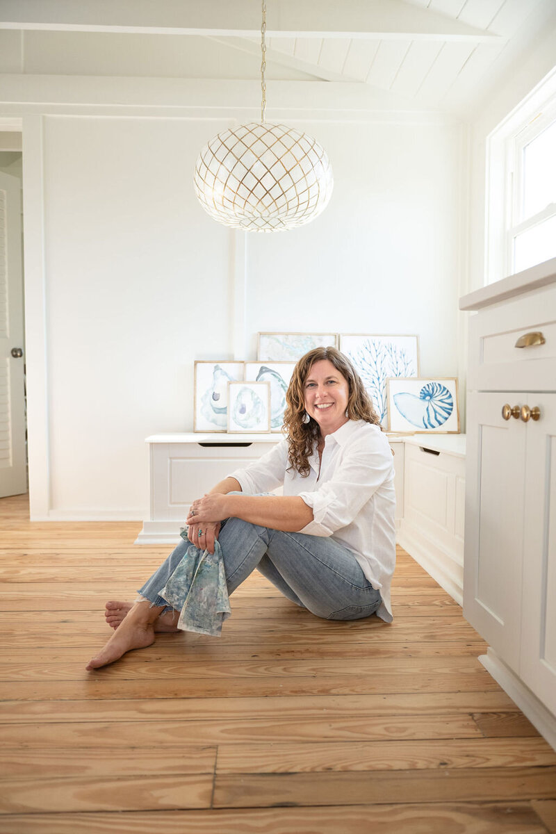 Laura, a watercolor artist sits on the wooden kitchen floor smiling with her arms crossed, a white globe chandelier overhead with her coastal watercolor artwork in the background