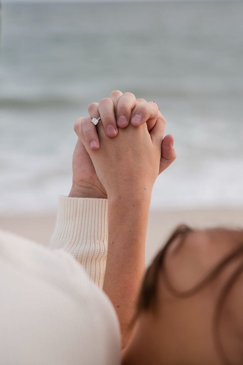 Couple with fingers entertwined, showing off the engagement ring with the ocean in the background.
