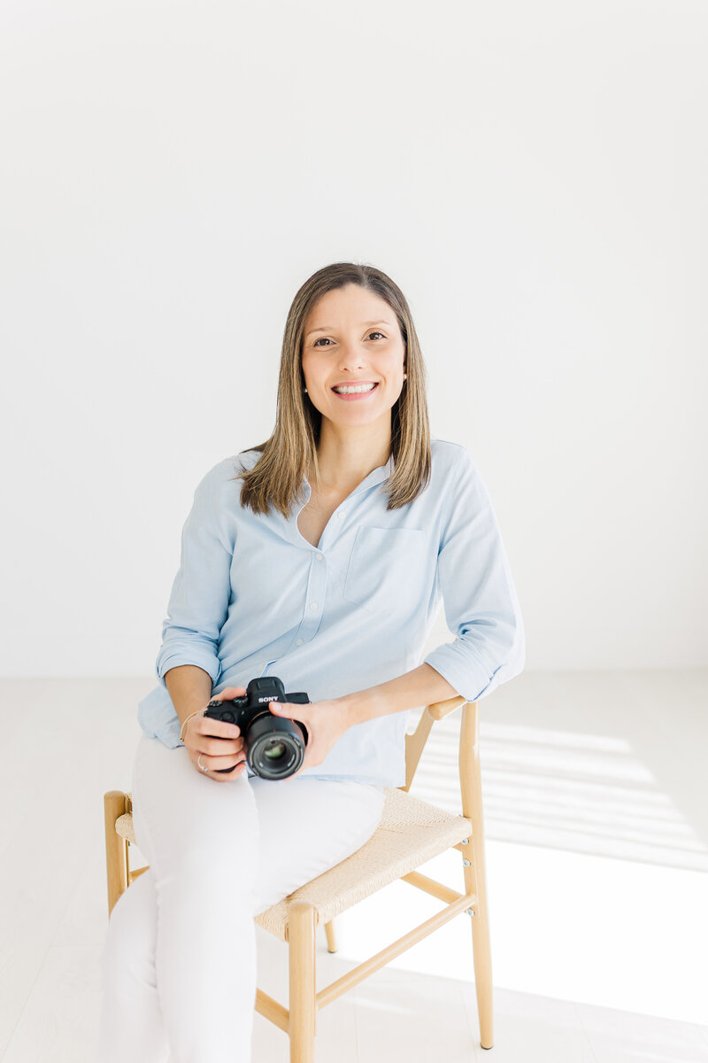 Headshot of an Austin Texas Photographer in a white studio. The photographer is dressed in a blue color shirt and white pants and leans on a chair. She smiles while looking at the camera.