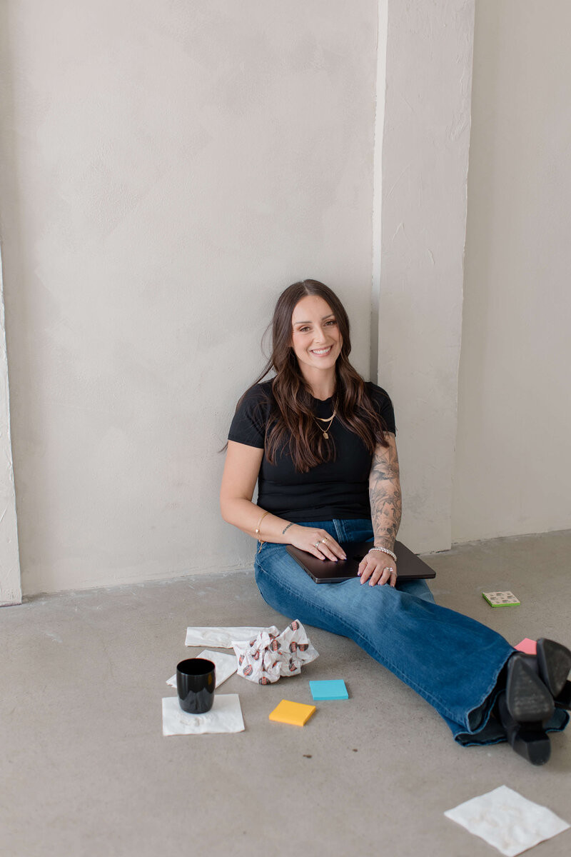 Sharon is sitting on the floor with a closed laptop in her lap and has some sticky notes around her smiling and posing with feet out ankles crossed.