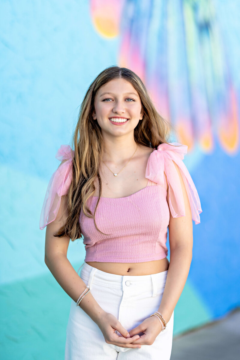 Senior girl smiling confidently in pink top during downtown session in Winter Garden, Florida by blue and pink wall.