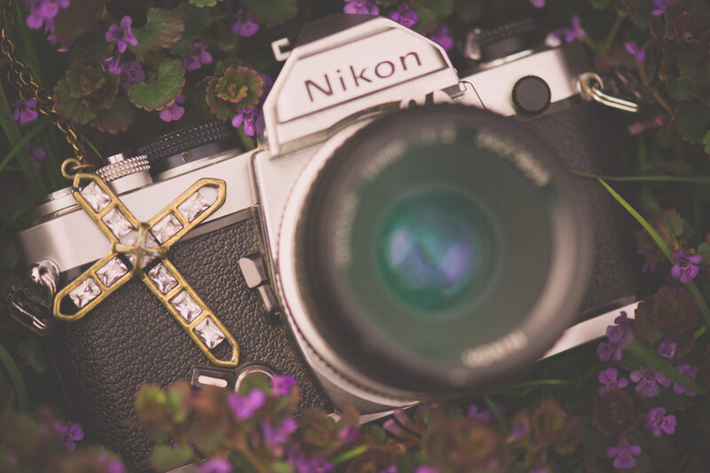 A Nikon film camera laying in purple flowers and grass with gold and diamond cross necklace by Kristin Palkoner.