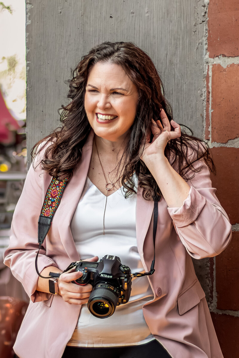 Branding photographer Jennifer Preiss holding camera and smiling in pink jacket against brick backdrop