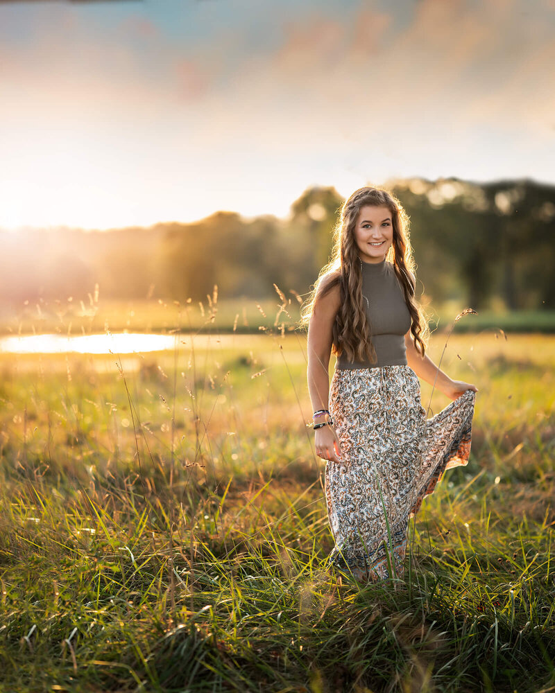 A Young girl in a field at sunset holds her skirt and twirls with sunlight in her hair. 