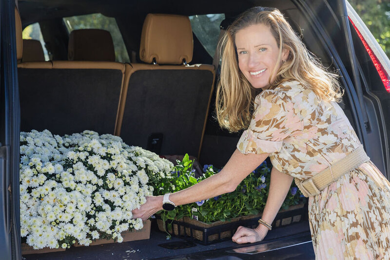 Woman with light brown blonde hair wearing light pink and brown floral dress smiling as she places plants and flowers in trunk of small SUV