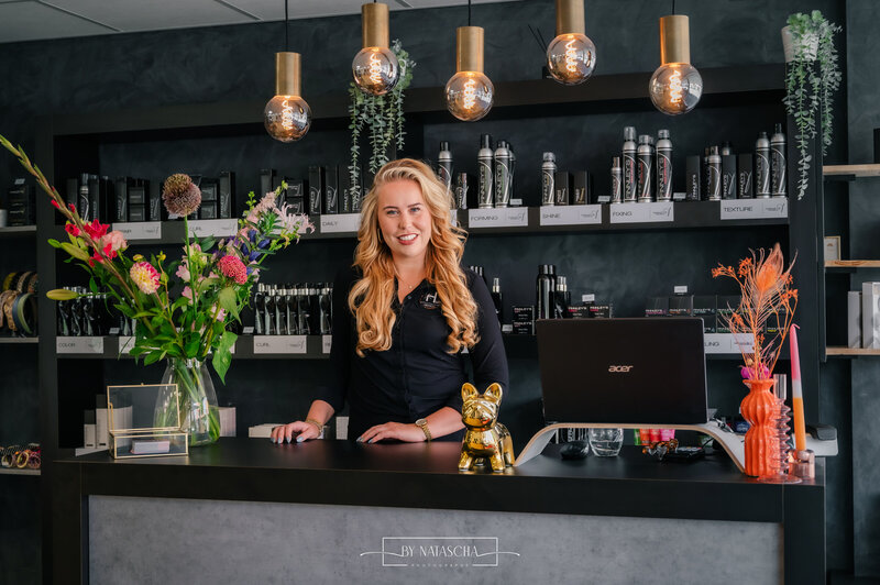 De eigenaar van de haarstudio bij de balie en kijkt lachend naar de camera