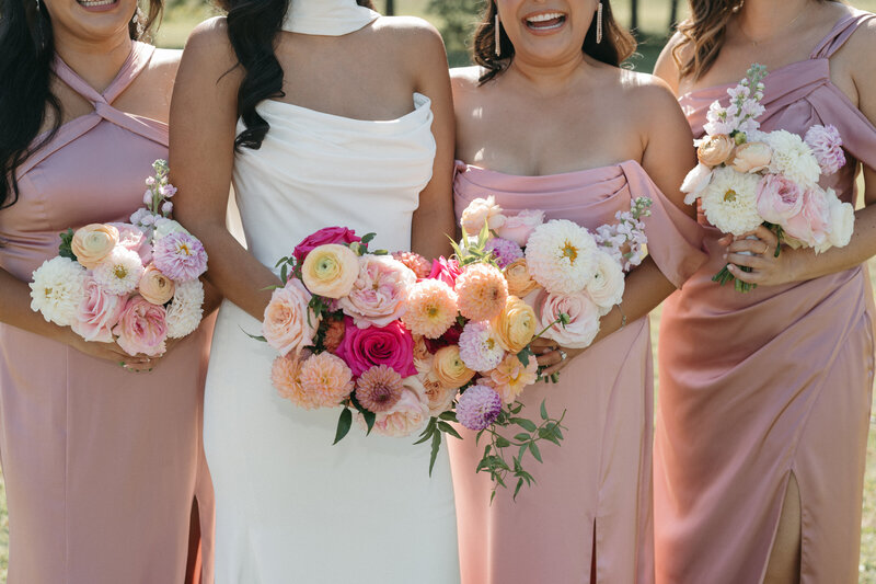 Close-up of vibrant garden-style wedding bouquets with dahlias, roses, ranunculus, and pastel blooms in peach, pink, and ivory hues, held by bride and bridesmaids in blush gowns—romantic floral styling.