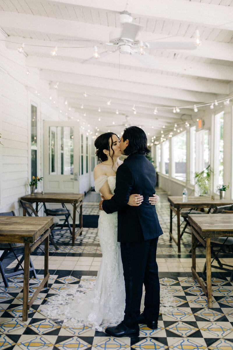 Hudson Valley bride and groom kiss on the porch under white ceiling, by lots of windows, on a bold graphic-tiled floor. Bride wears romantic lace dress.