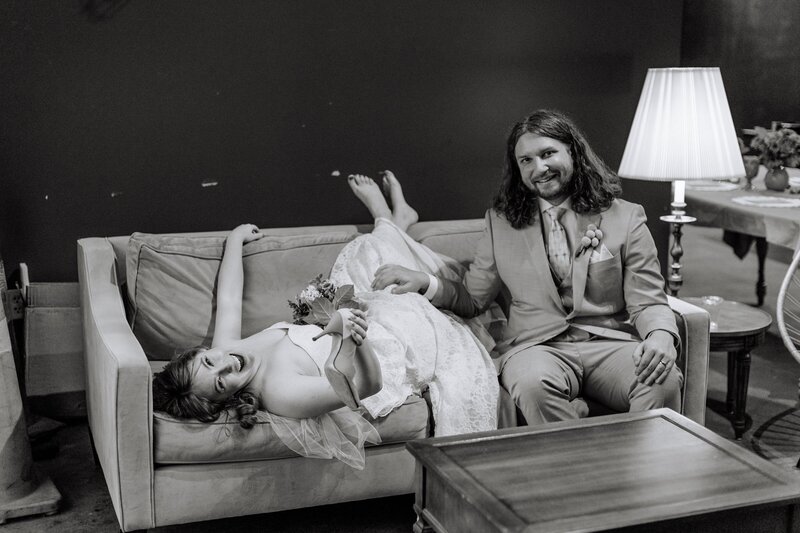 A bride laying on a couch in her wedding gown, laughing, with the groom sitting next to her with his hand on her leg