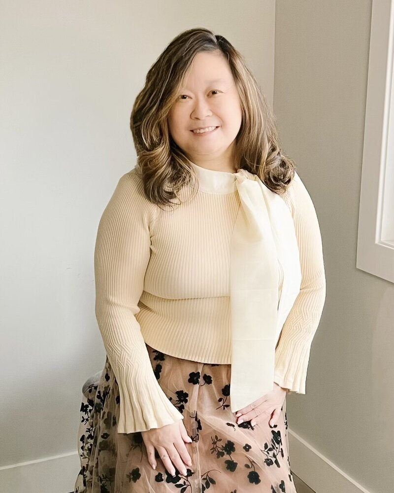 Anne, an Asian woman, is wearing a fine knit sweater with tie detail and a champagne tulle skirt with black velvet flower overlay detail. She is smiling at the camera.