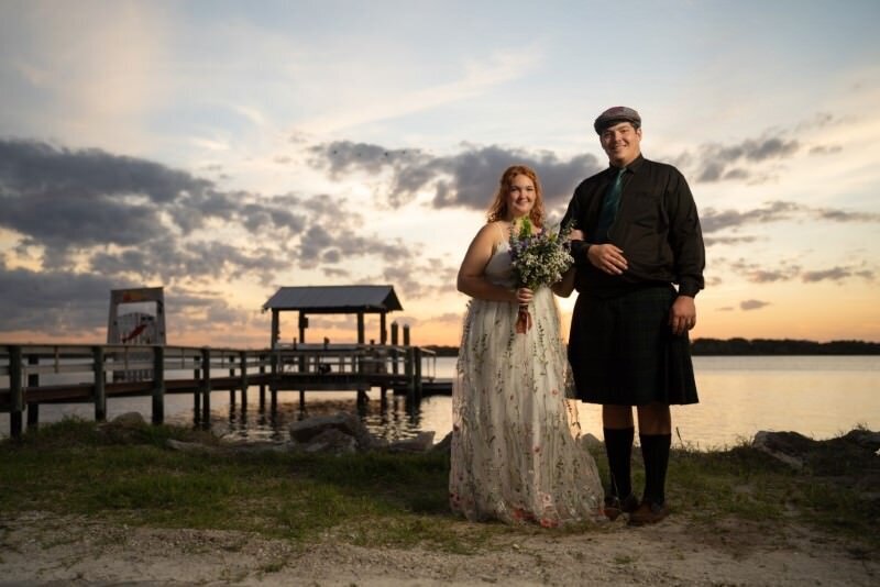 a bride in her white wedding dress holding a bouquet and a groom in a traditional Scottish kilt pose at sunset in front of water on their wedding day