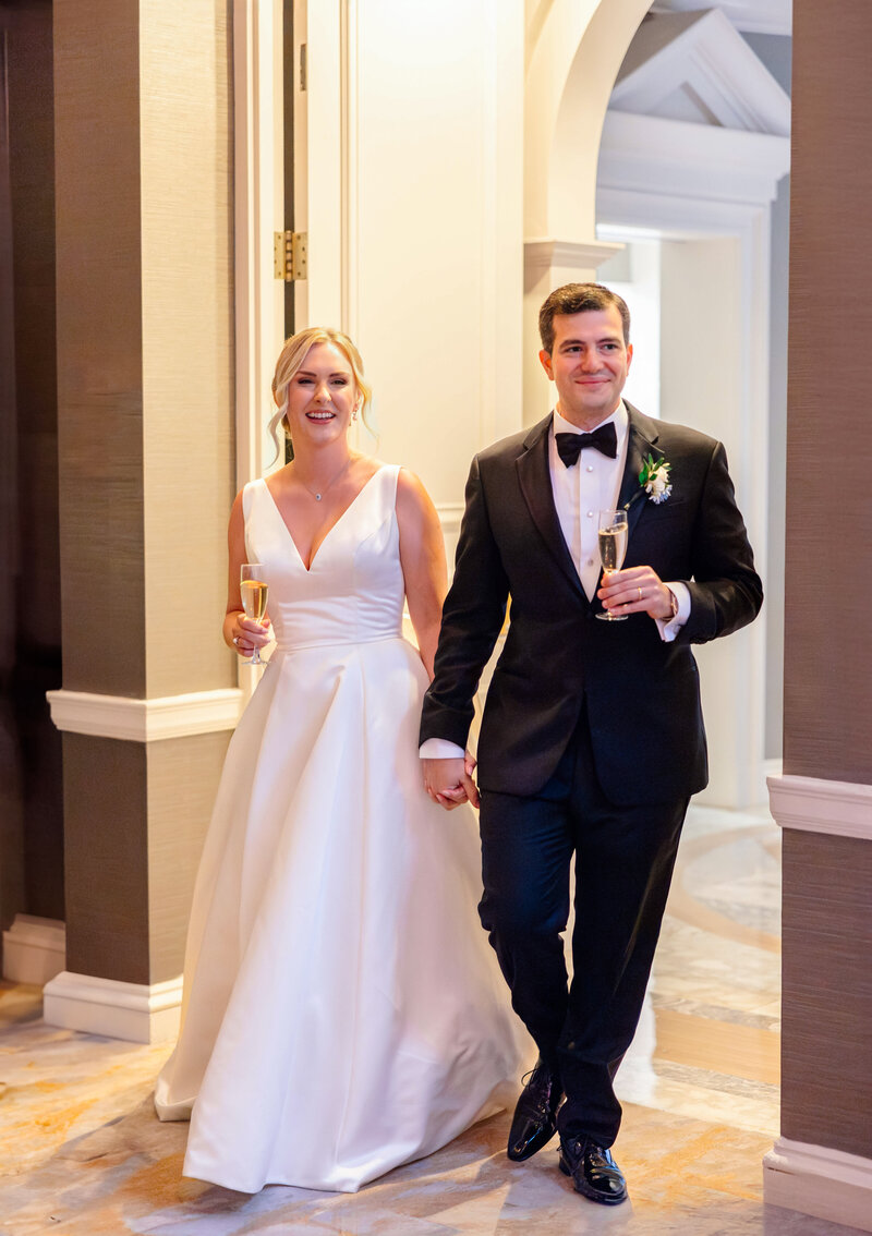 A couple in formal wedding attire walks hand in hand. The bride wears a white gown and holds a champagne flute. The groom is in a black tuxedo with a bow tie, also holding a glass. They appear to be smiling as they enter a room.