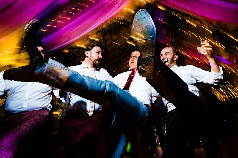 A groom with his father and groomsmen dancing at the reception kicking their legs in the air