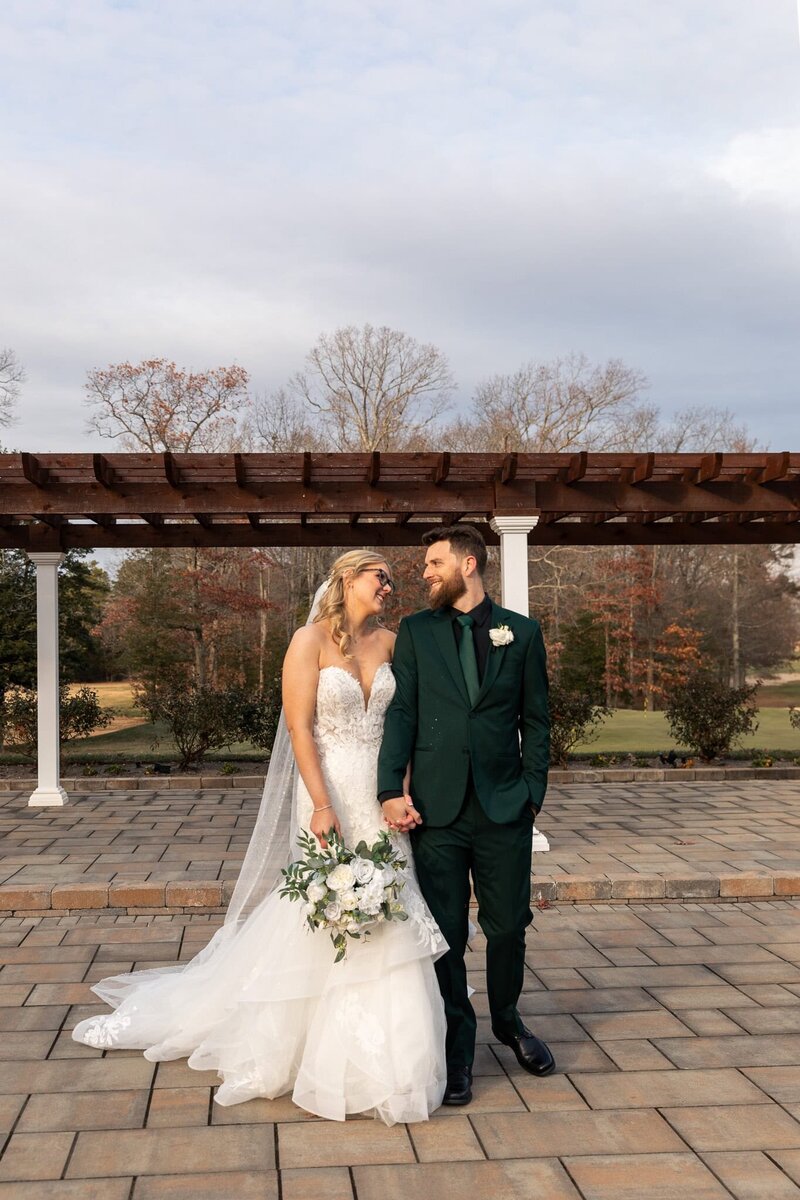 The bride and groom walk hand in hand across a stone patio, their smiles reflecting pure happiness. Her white lace wedding dress billows slightly as she holds a bouquet of white and green florals, while his deep green suit is accented with a classic boutonniere. Behind them, a wooden pergola and bare trees hint at a crisp Pennsylvania wedding day