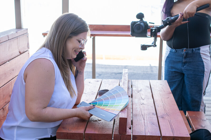 Smiling woman reviewing a color swatch book while talking on the phone at an outdoor wooden table, with a videographer filming nearby, photographed by Vyrl Photo to showcase Tucson brand photography.