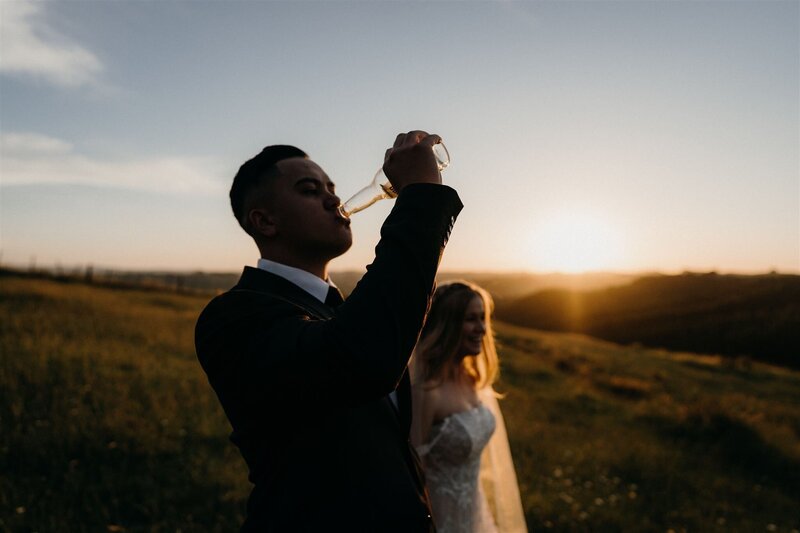 A groom drinking a beer in a wedding photo at sunset with the bride in the background photographed by tashina narelle, an auckland wedding photographer