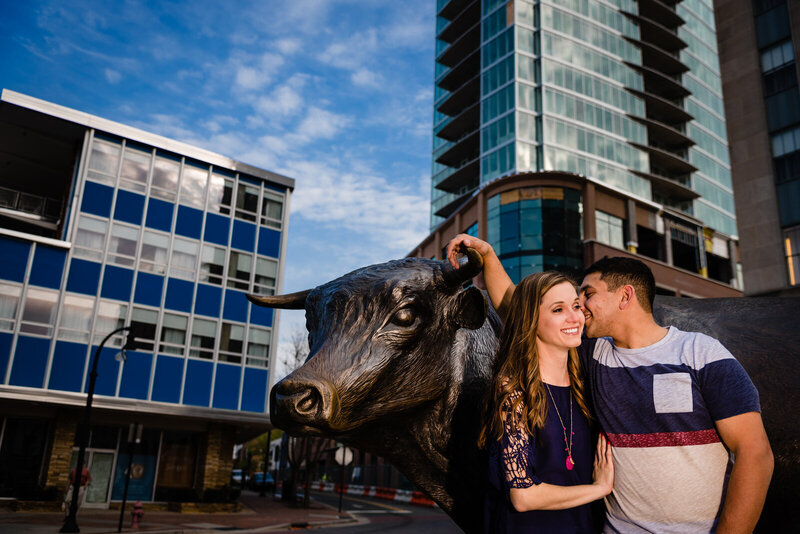 Man and woman pose with Major the Bull in downtown Durham, NC for the engagement photo session