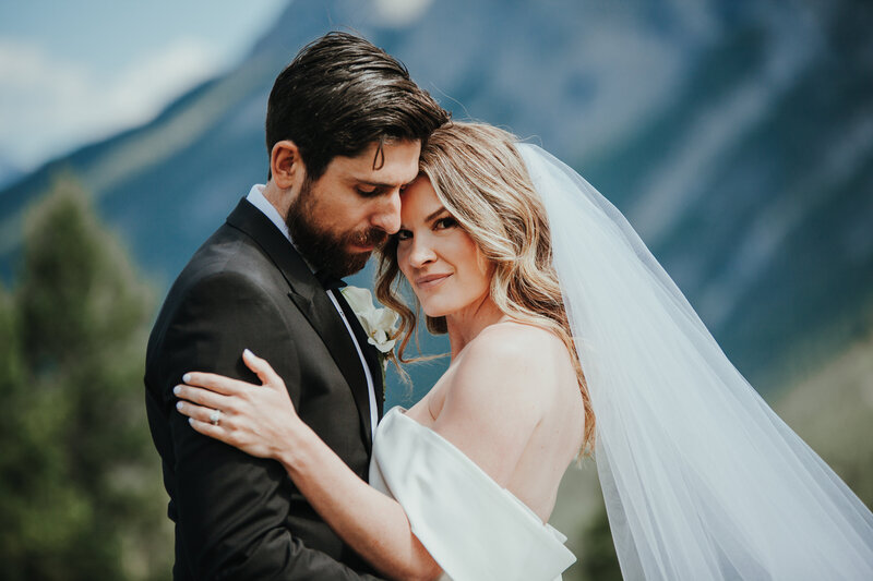 A couple who are eloping standi with their arms around each other face-to-face in front of a mountain range. The Bride looks directly at the camera smiling softly while the groom looks down on her with his arms around her waist.