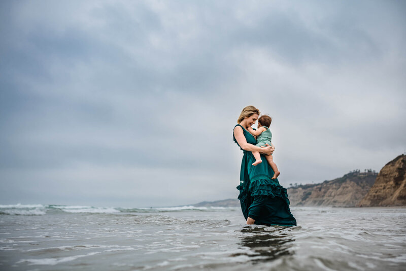 This is a photograph of a mother and her infant son while she holds him standing in the Pacific Ocean in La Jolla Shores. Photographed by Kim Belverud, a San Diego family photographer.